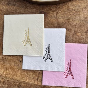 May include: Three square cocktail napkins in cream, white, and pink, each with an Eiffel Tower illustration and the phrase "She said Oui!" in a script font. The napkins are on a wooden surface.