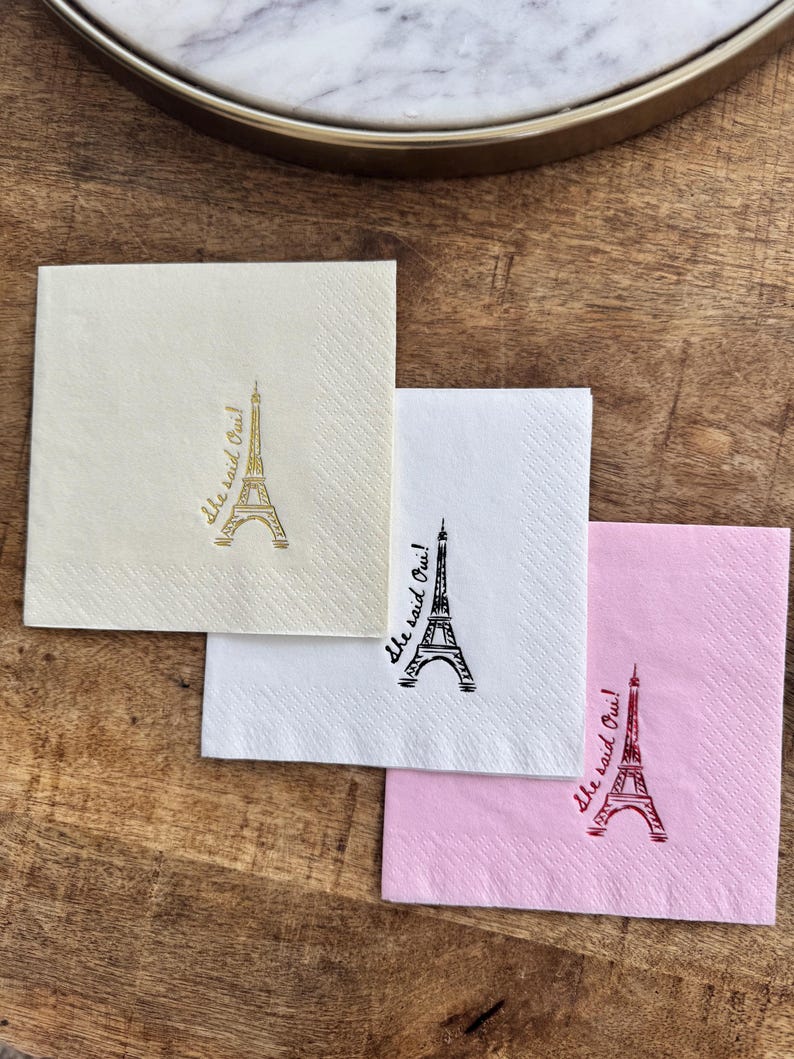 May include: Three square cocktail napkins in cream, white, and pink, each with an Eiffel Tower illustration and the phrase "She said Oui!" in a script font. The napkins are on a wooden surface.