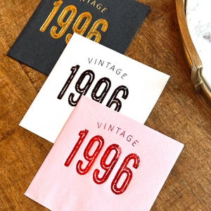 May include: Three square paper napkins with the word "VINTAGE" above the year "1996". One napkin is black with gold lettering, one is white with black lettering, and one is pink with red lettering.