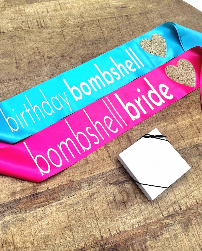 2 sashes the first one read birthday bombshell in white text with glitter gold heart on a turquoise sash.  The second sash reads bombshell bride with a glitter gold heart.  There is also a gift box in the picture with a bow which is included with the sash.