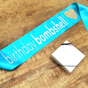 Op de afbeelding: Een turquoise satijnen sjerp met de woorden "birthday bombshell" in witte letters en een gouden glitterhart. Een stapel witte vierkante kaarten is vastgebonden met een zwart lint. Op een houten oppervlak.