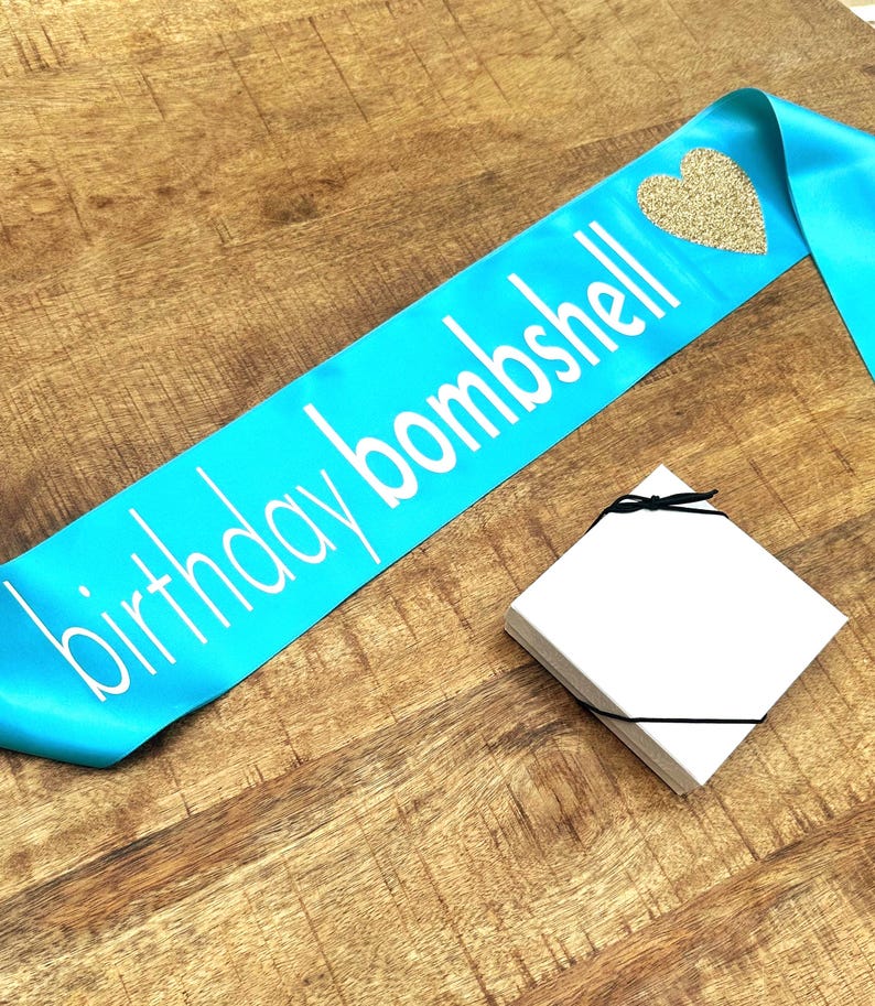 Op de afbeelding: Een turquoise satijnen sjerp met de woorden "birthday bombshell" in witte letters en een gouden glitterhart. Een stapel witte vierkante kaarten is vastgebonden met een zwart lint. Op een houten oppervlak.