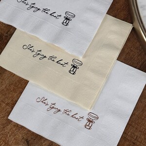 Three folded square napkins, stacked, with the top two in white and the bottom one in cream, each printed with "She's Tying the Knot" and a small bow illustration.