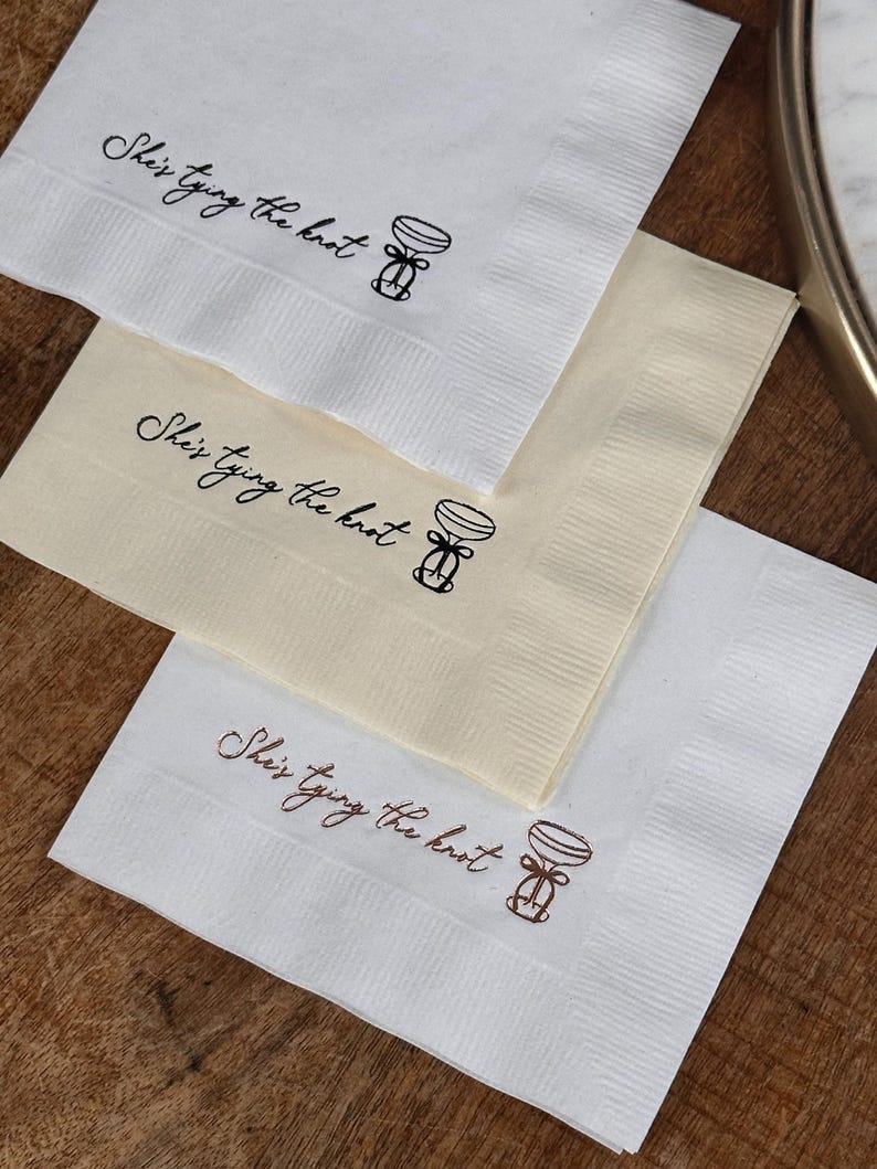 Three folded square napkins, stacked, with the top two in white and the bottom one in cream, each printed with "She's Tying the Knot" and a small bow illustration.