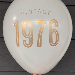 Puede incluir: Globo blanco con la palabra "VINTAGE" y el n&uacute;mero "1976" impresos en dorado. El globo es sostenido por una mano. El fondo es gris oscuro. Adecuado para una celebraci&oacute;n de cumplea&ntilde;os o aniversario.