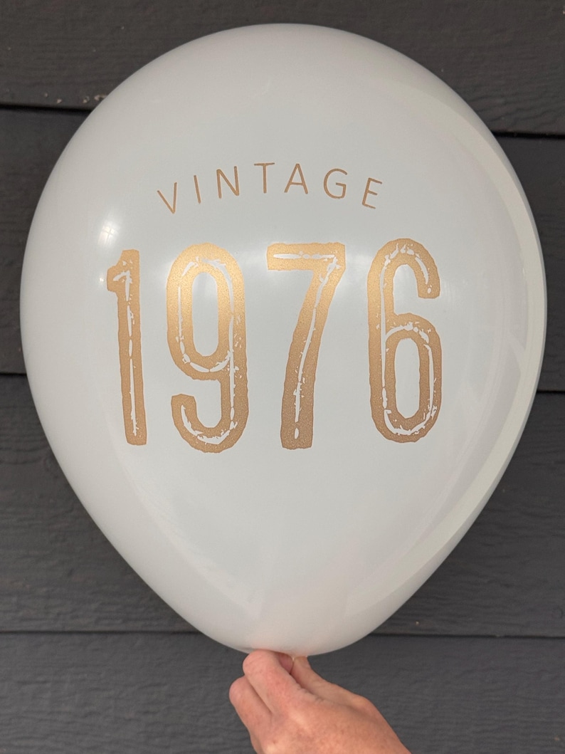 Puede incluir: Globo blanco con la palabra "VINTAGE" y el n&uacute;mero "1976" impresos en dorado. El globo es sostenido por una mano. El fondo es gris oscuro. Adecuado para una celebraci&oacute;n de cumplea&ntilde;os o aniversario.