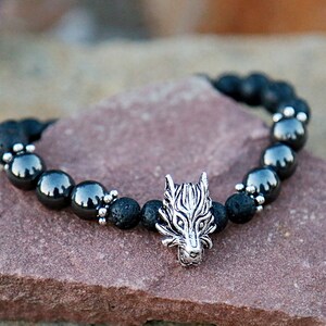 Pode incluir: Uma pulseira de contas pretas com um pingente de cabe&ccedil;a de lobo prateado. A pulseira &eacute; feita de rocha vulc&acirc;nica preta e contas de hematita cinza escuro. O pingente de cabe&ccedil;a de lobo &eacute; detalhado com linhas gravadas.