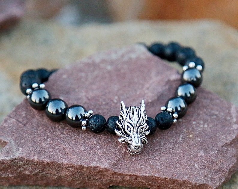 Pode incluir: Uma pulseira de contas pretas com um pingente de cabe&ccedil;a de lobo prateado. A pulseira &eacute; feita de rocha vulc&acirc;nica preta e contas de hematita cinza escuro. O pingente de cabe&ccedil;a de lobo &eacute; detalhado com linhas gravadas.