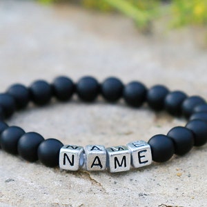 Puede incluir: Pulsera de cuentas negras con cuentas de letras plateadas que deletrean la palabra "NAME".