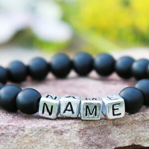 Puede incluir: Un brazalete de cuentas negras con cuentas de letras plateadas que deletrean la palabra "NAME".