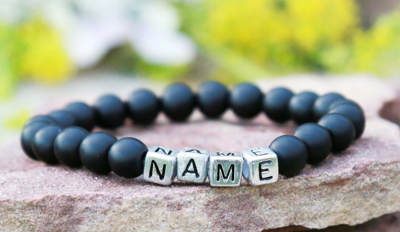 Puede incluir: Un brazalete de cuentas negras con cuentas de letras plateadas que deletrean la palabra "NAME".