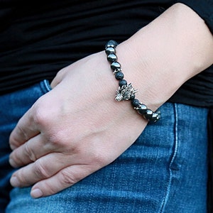 Pode incluir: Uma pulseira de contas com pedras de lava pretas e contas de hematita cinza escuro, com um pingente de cabe&ccedil;a de drag&atilde;o prateado. A pulseira &eacute; usada no pulso. Fundo preto e jeans azuis.