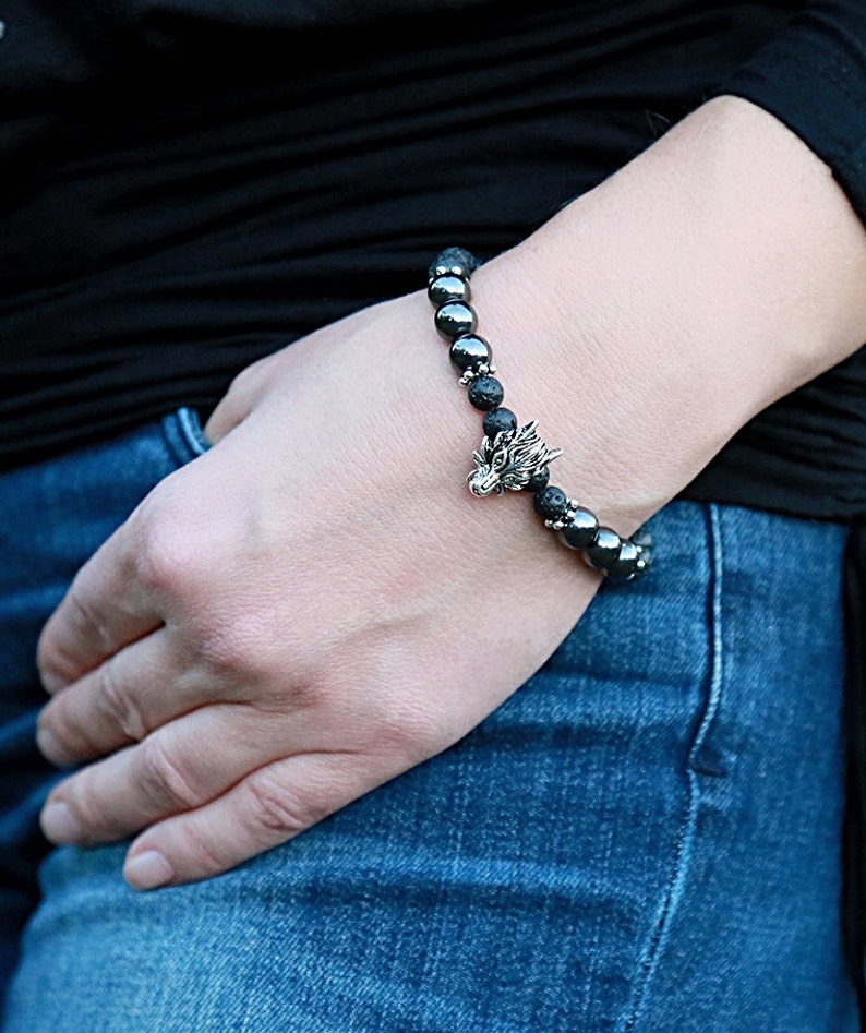 Pode incluir: Uma pulseira de contas com pedras de lava pretas e contas de hematita cinza escuro, com um pingente de cabe&ccedil;a de drag&atilde;o prateado. A pulseira &eacute; usada no pulso. Fundo preto e jeans azuis.