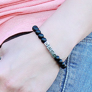 Puede incluir: Un brazalete de cuentas negras con cuentas plateadas que deletrean la palabra "NAME".
