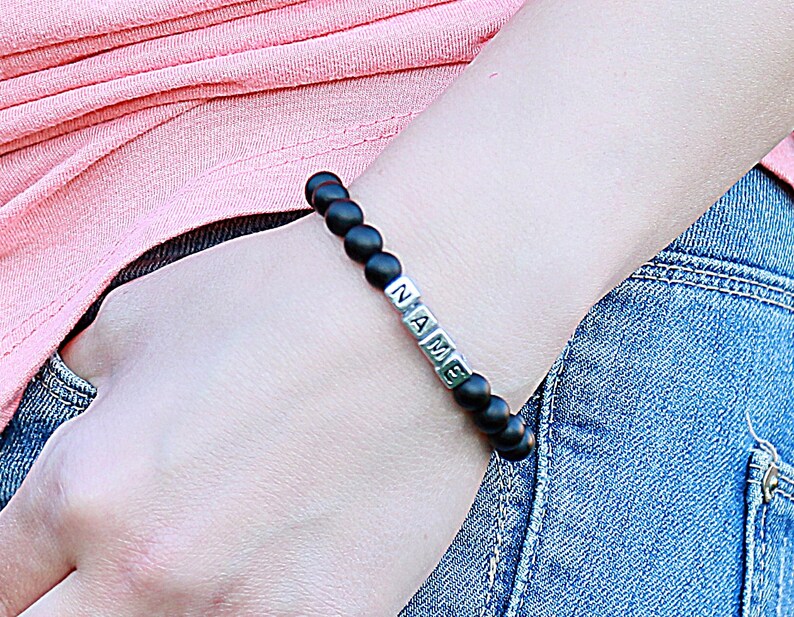 Puede incluir: Un brazalete de cuentas negras con cuentas plateadas que deletrean la palabra "NAME".