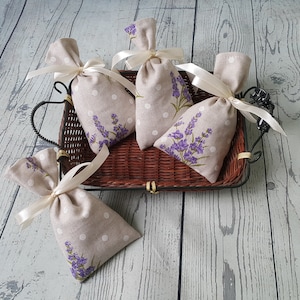 以下が含まれることがあります： 白のドットとラベンダーの花柄が施された、ベージュのリネン製の小さな袋が5つ。袋は白いサテンリボンで結ばれており、茶色の籐のバスケットに並べられています。