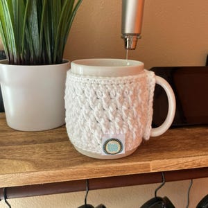 May include: A white ceramic mug with a white crocheted cosy. The cosy has a textured pattern and a small label. A silver frother dispenses liquid into the mug. A potted plant sits to the left on a wooden shelf. Black mugs hang below.