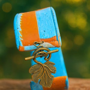 May include: A handcrafted bracelet with a unique design. The bracelet features a blue and orange painted wooden base, accented by a bronze-colored leaf charm. The charm is suspended from a decorative clasp. The bracelet is a stylish accessory.
