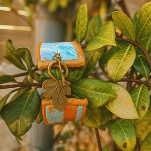 May include: A decorative keychain featuring two small, rectangular boxes with blue and orange painted details. The boxes are connected by a brass key and a leaf-shaped charm. The background includes green leaves and branches.