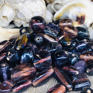 May include: A collection of iridescent glass beads in shades of purple and black. The beads are various shapes and sizes, including round, oval, and rectangular.