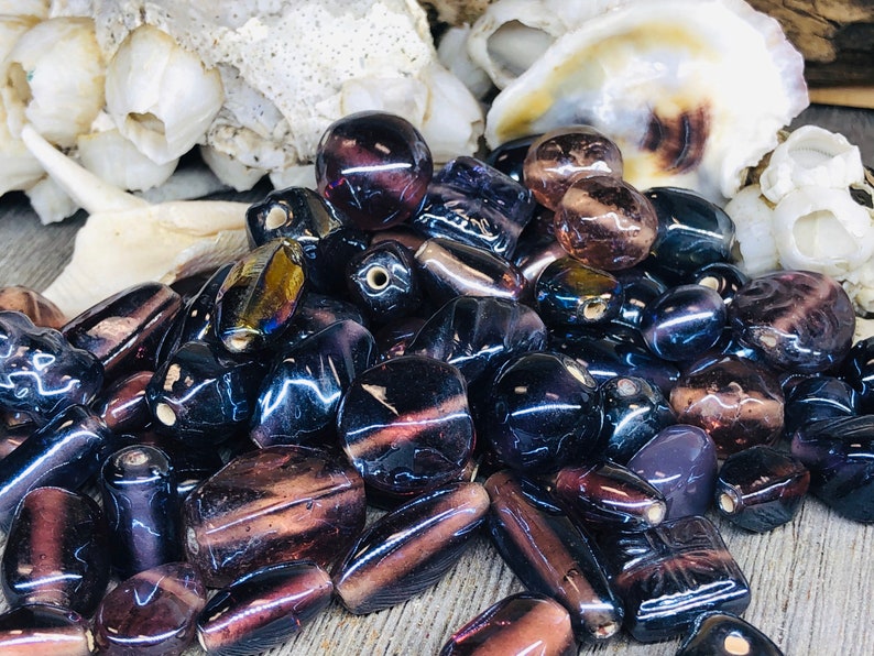 May include: A collection of iridescent glass beads in shades of purple and black. The beads are various shapes and sizes, including round, oval, and rectangular.