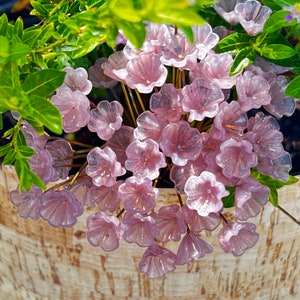 以下が含まれることがあります： 花瓶にアレンジされた、繊細で透き通ったピンク色のガラスの花のブーケ。花はガラス製で、繊細で女性らしい印象です。