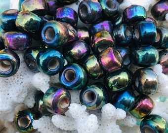 May include: A collection of iridescent, barrel-shaped beads. The beads have a dark base color with a shimmering, multicolored finish that reflects hues of blue, purple, and green. They are arranged on a white, textured surface.