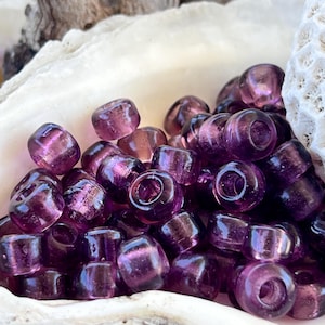Op de afbeelding: Een verzameling doorschijnende paarse glazen kralen met een cilindrische vorm. De kralen hebben een gat in het midden en worden gepresenteerd in een witte zeeschelp. De kralen hebben een glanzende afwerking.