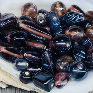 May include: A collection of dark purple and black glass beads of various shapes and sizes. The beads are translucent and have a glossy finish.