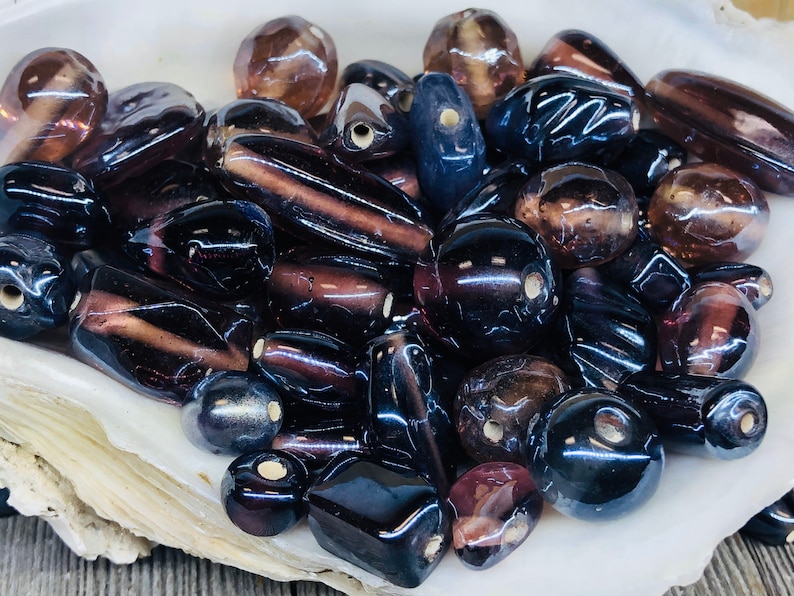 May include: A collection of dark purple and black glass beads of various shapes and sizes. The beads are translucent and have a glossy finish.