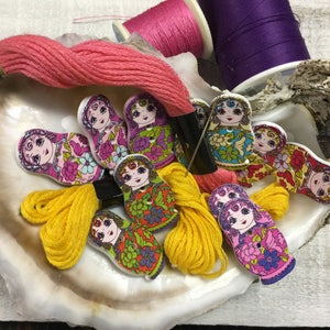 May include: A collection of colourful wooden nesting doll buttons with floral patterns. The buttons are arranged in a seashell with pink, yellow, and purple embroidery floss.