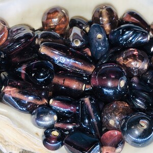 May include: A collection of dark purple and brown glass beads in various shapes and sizes. The beads have a shiny, iridescent finish.