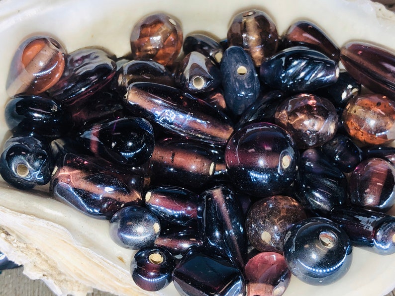 May include: A collection of dark purple and brown glass beads in various shapes and sizes. The beads have a shiny, iridescent finish.