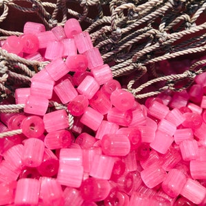 Op de afbeelding: Een close-up van een stapel roze glazen kralen. De kralen zijn cilindrisch en hebben een licht iriserende afwerking. Ze zijn in een willekeurig patroon gerangschikt en zijn gedeeltelijk bedekt met een stuk bruin touw.