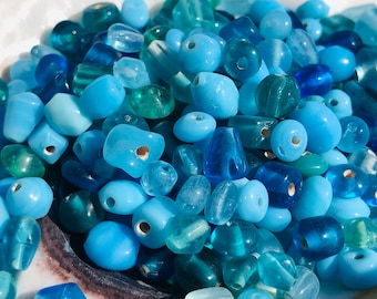Puede incluir: Una colección de cuentas de vidrio azules y verde azulado de varias formas y tamaños. Las cuentas son perfectas para hacer joyas o manualidades.