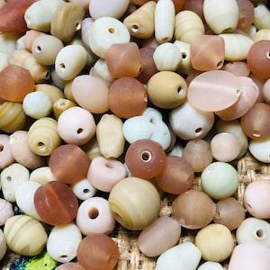May include: A collection of smooth, pastel-colored glass beads in various shapes and sizes. The beads are arranged in a random pattern and are mostly in shades of beige, brown, and white.