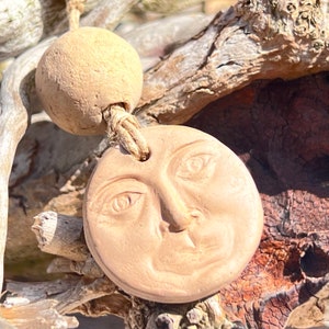 May include: A clay pendant with a face sculpted on it. The pendant is attached to a twine cord with a clay bead at the top.