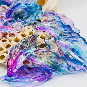 May include: A collection of iridescent glass leaf-shaped beads in shades of blue, purple, and gold. The beads are scattered on a textured, off-white surface, creating a shimmering effect. The glass has a translucent quality.