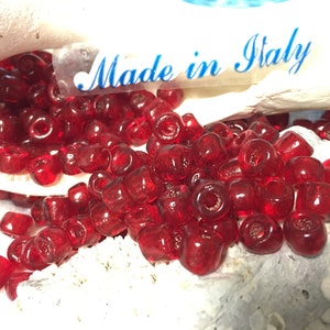 May include: A pile of small, red, glass beads. The beads are round with a hole in the centre. The beads are made in Italy.