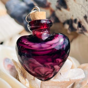 May include: A heart-shaped glass bottle with a cork stopper and a silver loop. The bottle contains a swirling mix of purple and pink hues. The bottle is set on a bed of seashells.