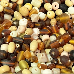 Puede incluir: Una colección de cuentas variadas en diversas formas y tamaños. Las cuentas son de color crema, marrón y ámbar, algunas con diseños a rayas o estampados. Estas cuentas, hechas de vidrio o material similar, son adecuadas para la creación de joyas.