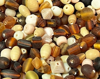 May include: A collection of assorted beads in various shapes and sizes. The beads are in shades of cream, brown, and amber, with some having striped or patterned designs. The beads are made of glass or a similar material and are suitable for jewelry making.