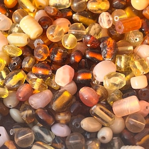 May include: A collection of colourful glass beads in various shapes and sizes, including round, oval, and rectangular. The beads are a mix of brown, amber, yellow, and pink.