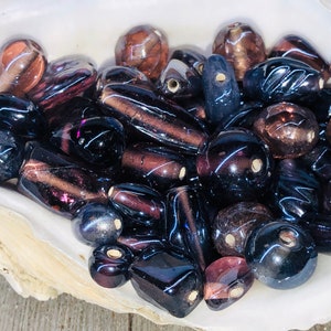 May include: A collection of iridescent glass beads in shades of purple, brown, and black. The beads are various shapes and sizes, including round, oval, and rectangular.