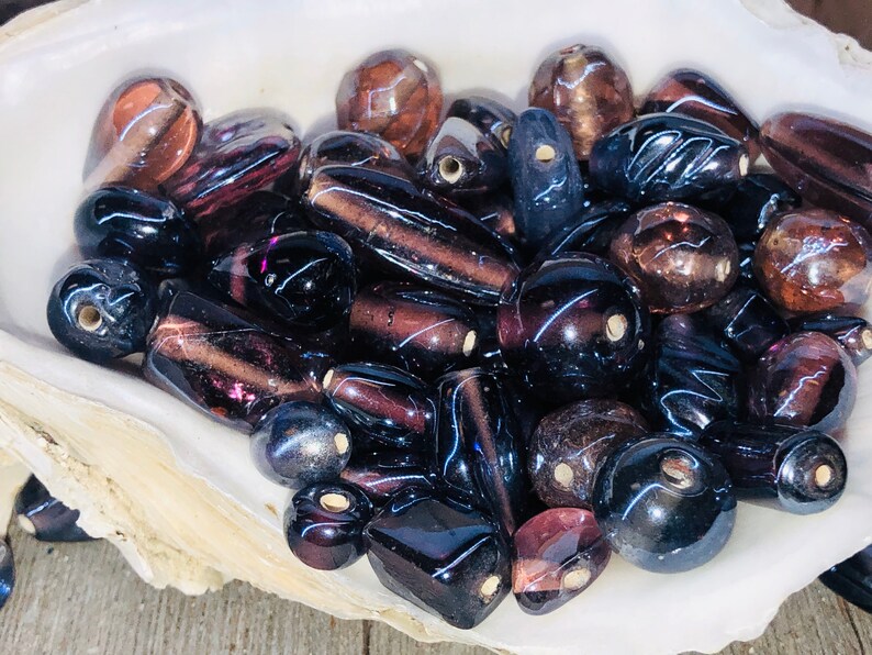 May include: A collection of iridescent glass beads in shades of purple, brown, and black. The beads are various shapes and sizes, including round, oval, and rectangular.