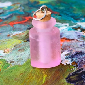 May include: A small pink glass bottle with a cork stopper and a silver metal loop. The bottle is sitting on a colorful, abstract background.