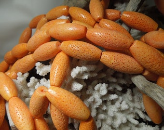 May include: A strand of orange coral beads, each bead is oval shaped and has a textured surface. The beads are arranged in a pile on a white coral background.