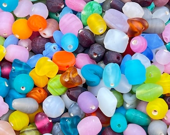 Puede incluir: Una colección de cuentas de vidrio de colores en varias formas y tamaños. Las cuentas son una mezcla de colores opacos y translúcidos, incluyendo azul, verde, rosa, morado y amarillo.