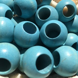 以下が含まれることがあります： 各ビーズの中心に穴が開いた、ターコイズブルーのウッドビーズの山をクローズアップで撮影したものです。ビーズは滑らかで光沢のある仕上げになっています。
