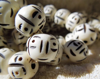 May include: Close-up of a cream-colored beaded bracelet. Each bead is round with black etched facial designs. The beads are strung together, creating a circular pattern. The bracelet is likely handmade and suitable for everyday wear.